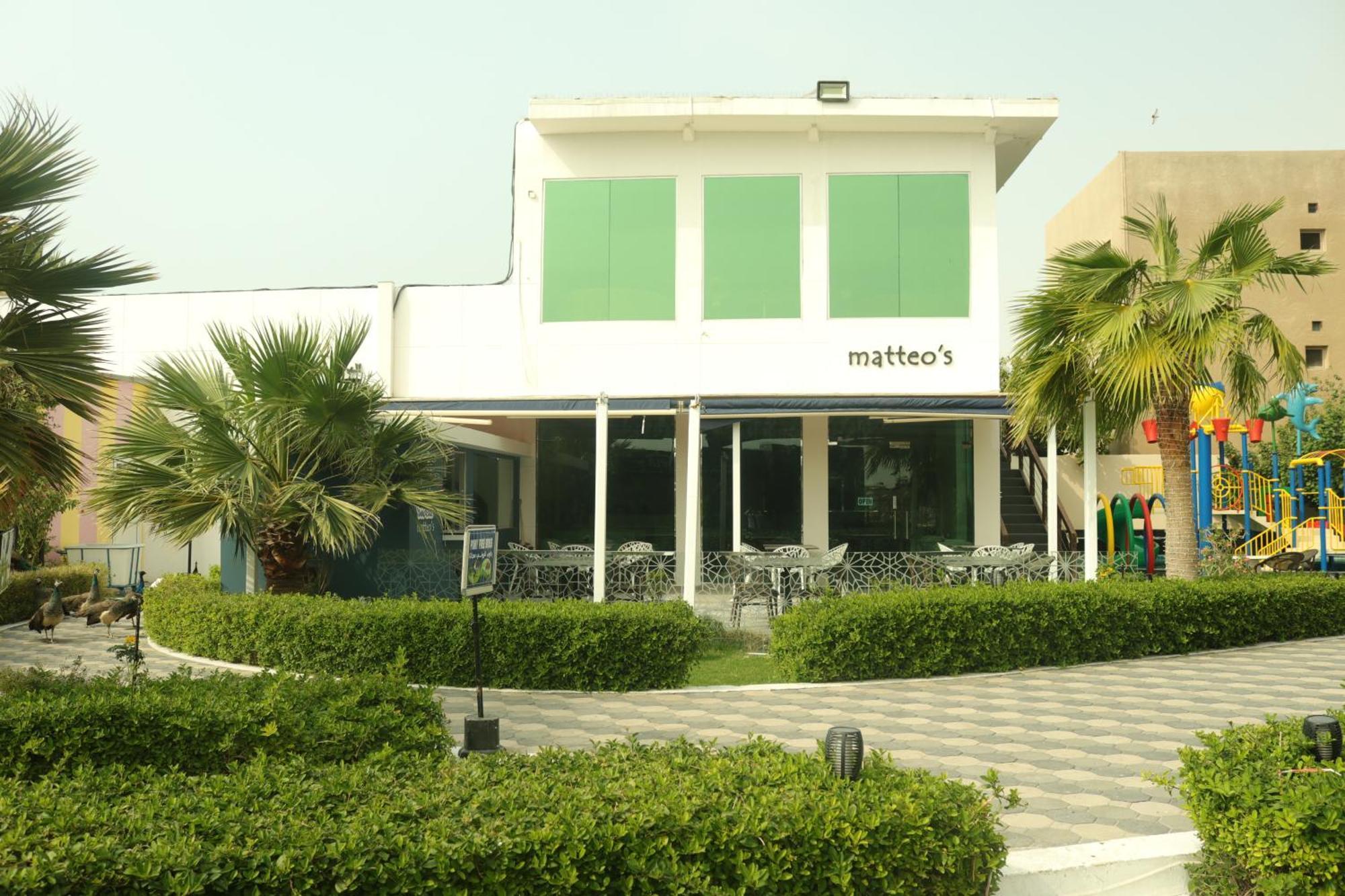 Lagoona Beach Luxury Resort And Spa Μανάμα Εξωτερικό φωτογραφία The photo shows a building with the name "matteo's" displayed prominently on the front. The structure features large windows and green shutters. There is a landscaped area in front with bushes and palm trees, along with a paved pathway leading up to 