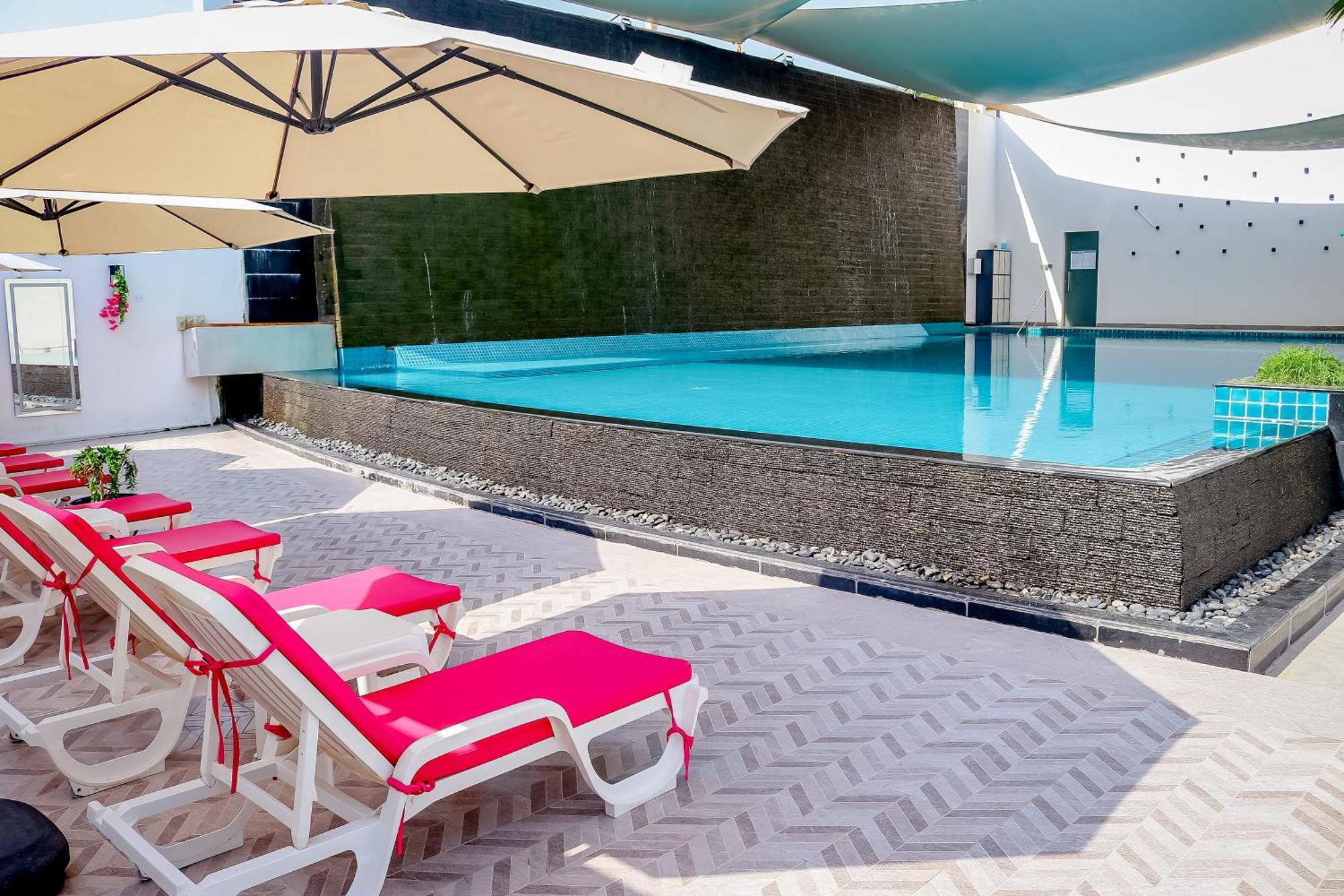 Lagoona Beach Luxury Resort And Spa Μανάμα Εξωτερικό φωτογραφία The photo shows a pool area featuring a rectangular swimming pool with clear blue water. Surrounding the pool are light-colored lounge chairs with pink cushions, positioned under large umbrellas for shade. The pool is bordered by a textured wall, and