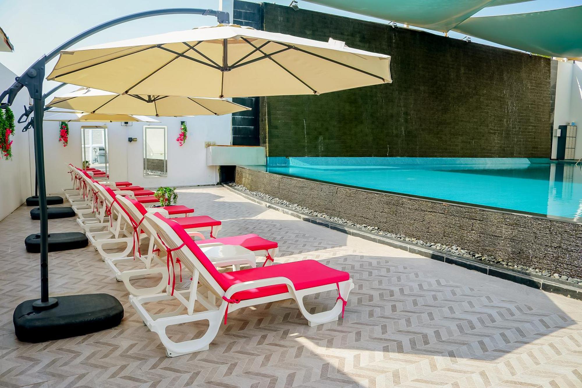 Lagoona Beach Luxury Resort And Spa Μανάμα Εξωτερικό φωτογραφία The photo shows a sunny poolside area with several lounge chairs arranged in a row. Each chair features red cushions and is positioned under large, beige umbrellas for shade. In the background, there is a beautiful swimming pool with clear blue water