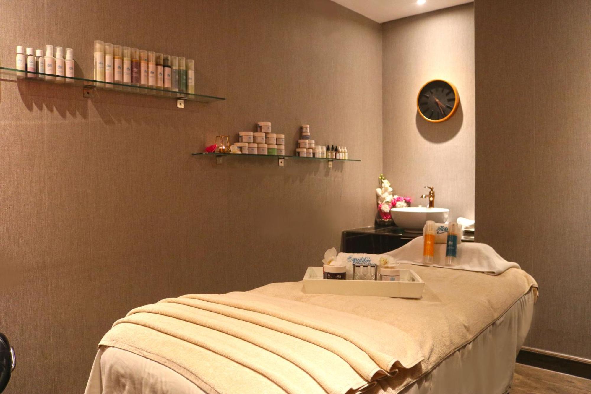 Lagoona Beach Luxury Resort And Spa Μανάμα Εξωτερικό φωτογραφία The photo shows a spa treatment room. There is a massage table draped with a light-colored blanket, creating a relaxing atmosphere. On the wall, there are shelves displaying various skincare products in bottles and jars. A clock is mounted on the wal