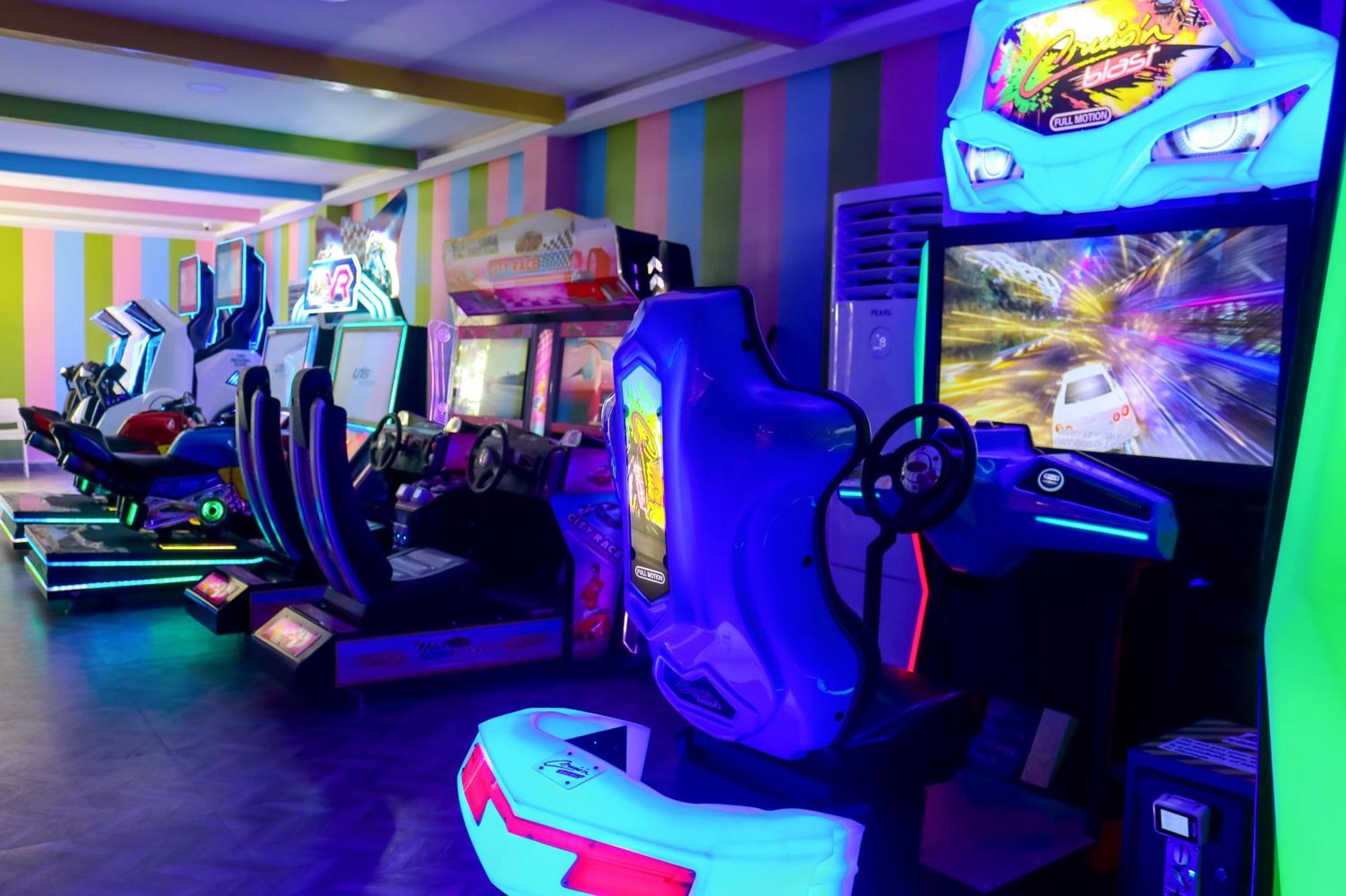 Lagoona Beach Luxury Resort And Spa Μανάμα Εξωτερικό φωτογραφία The photo shows an arcade area filled with various arcade machines. The setting is illuminated with colorful neon lights, creating a vibrant atmosphere. There are several racing game machines with steering wheels and seats, each accompanied by screen