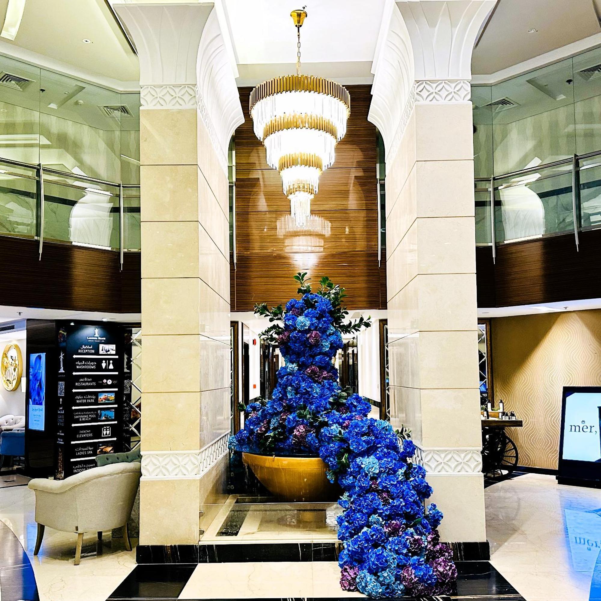 Lagoona Beach Luxury Resort And Spa Μανάμα Εξωτερικό φωτογραφία The photo depicts an elegant lobby or entrance area of a building. Central to the scene is a large floral arrangement featuring vibrant blue flowers. This arrangement is positioned in a decorative bowl. Above it hangs a stylish chandelier with a laye