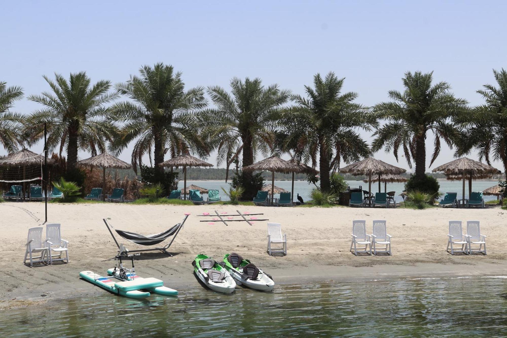 Lagoona Beach Luxury Resort And Spa Μανάμα Εξωτερικό φωτογραφία The photo depicts a serene beach scene with several palm trees lining the shore. There are thatched umbrellas providing shade, and a few lounge chairs are positioned on the sand. A hammock is set up between two supports, inviting relaxation. In the f