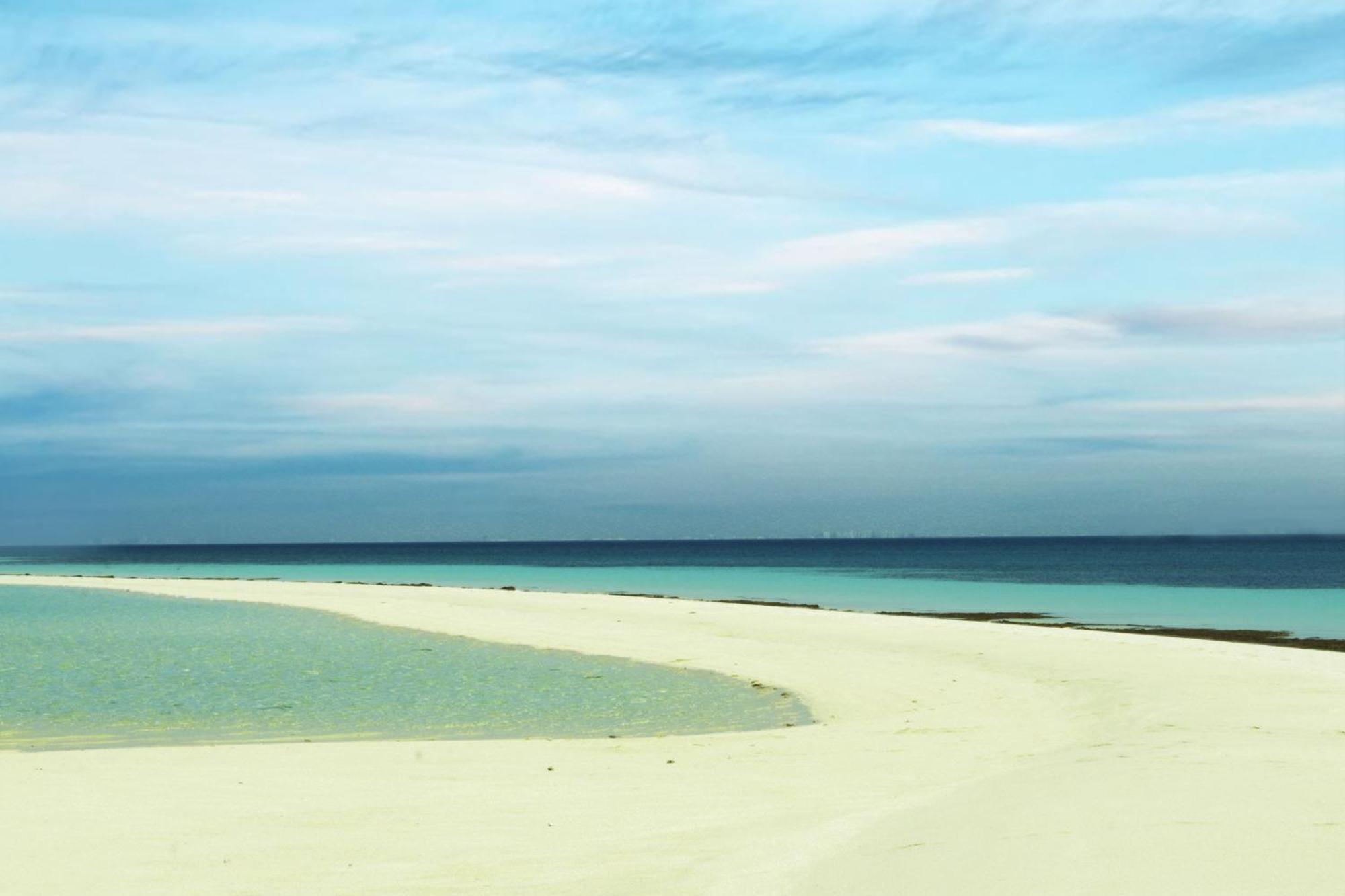 Lagoona Beach Luxury Resort And Spa Μανάμα Εξωτερικό φωτογραφία The photo depicts a serene beach scene with a gentle curve of sandy shore leading into shallow turquoise waters. The calm ocean stretches into the horizon, meeting a sky filled with soft clouds in various shades of blue. The overall atmosphere is tra