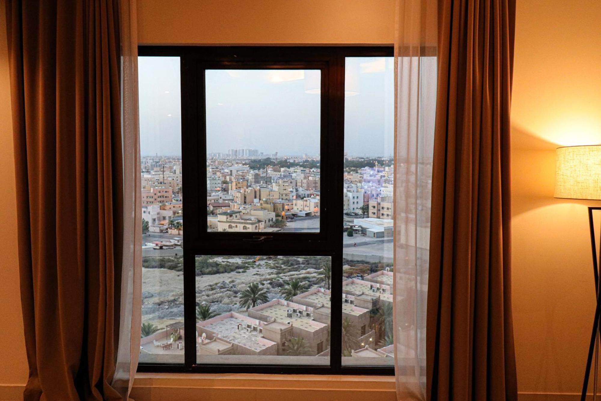 Lagoona Beach Luxury Resort And Spa Μανάμα Εξωτερικό φωτογραφία The photo shows a window with a black frame and sheer curtains. Through the window, there is a view of a cityscape, displaying a densely populated area with various buildings and rooftops. The sky appears slightly overcast, suggesting a muted lightin