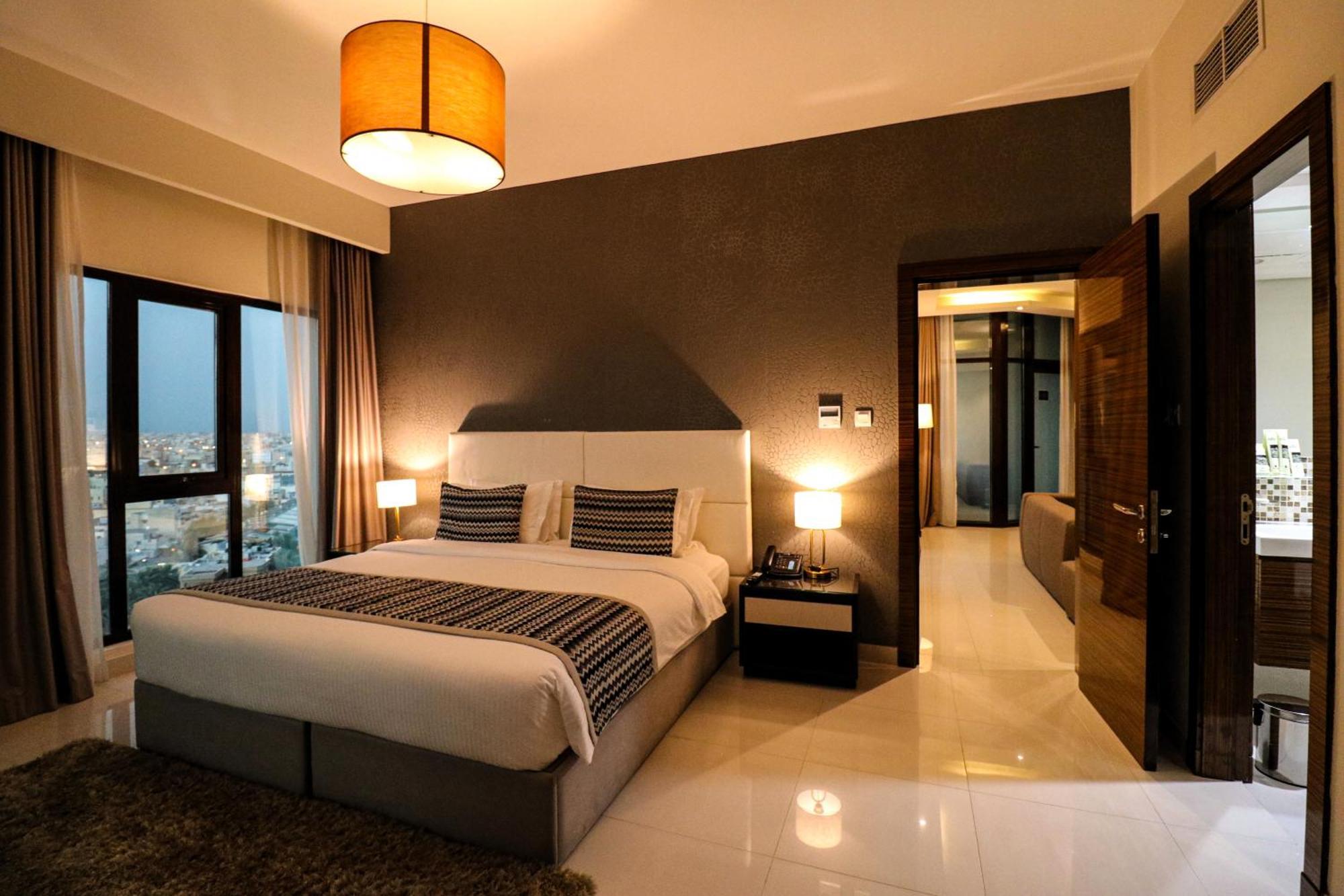 Lagoona Beach Luxury Resort And Spa Μανάμα Εξωτερικό φωτογραφία The photo depicts a modern hotel room or suite. It features a large bed with a white and gray color scheme, accented with decorative pillows. On either side of the bed, there are sleek bedside lamps providing warm lighting. The walls are a dark color