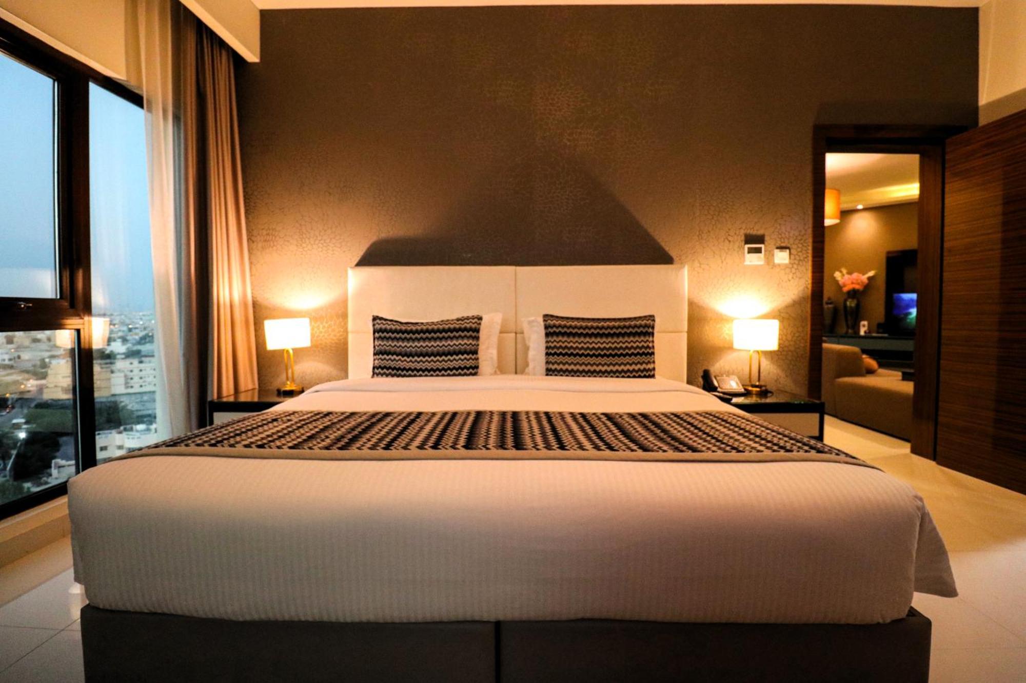 Lagoona Beach Luxury Resort And Spa Μανάμα Εξωτερικό φωτογραφία The photo depicts a modern hotel room featuring a large bed with a white comforter and two decorative pillows. The headboard is light-colored, and the walls are painted a dark hue. On either side of the bed, there are bedside tables with lamps. The r