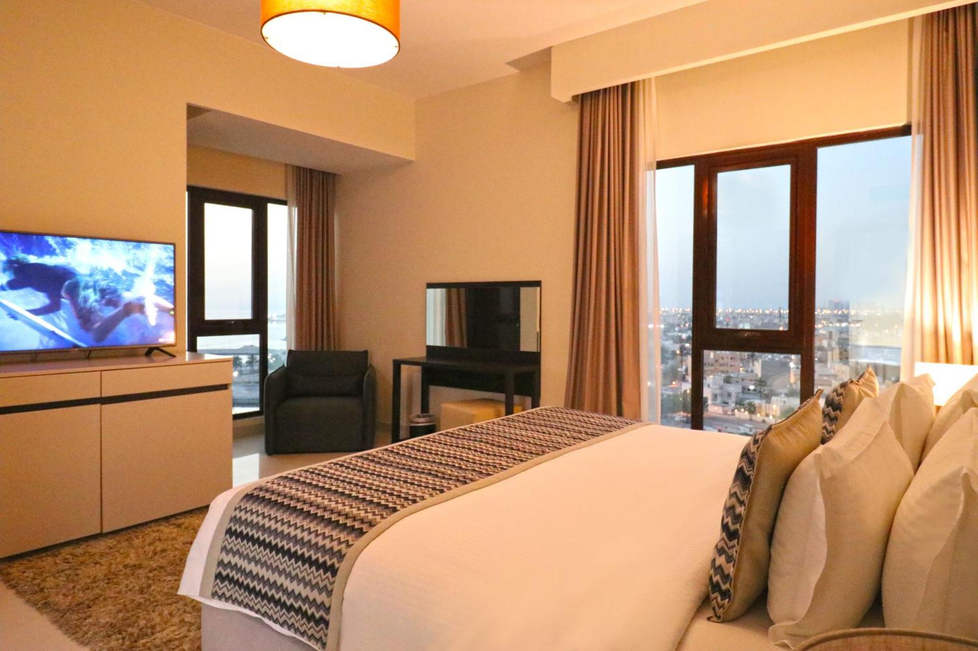 Lagoona Beach Luxury Resort And Spa Μανάμα Εξωτερικό φωτογραφία The photo shows a modern hotel room or suite. It features a neatly made bed with decorative pillows in the foreground. To the left, there is a TV on a cabinet, and further back, a comfortable chair is situated in front of a large window that likely o
