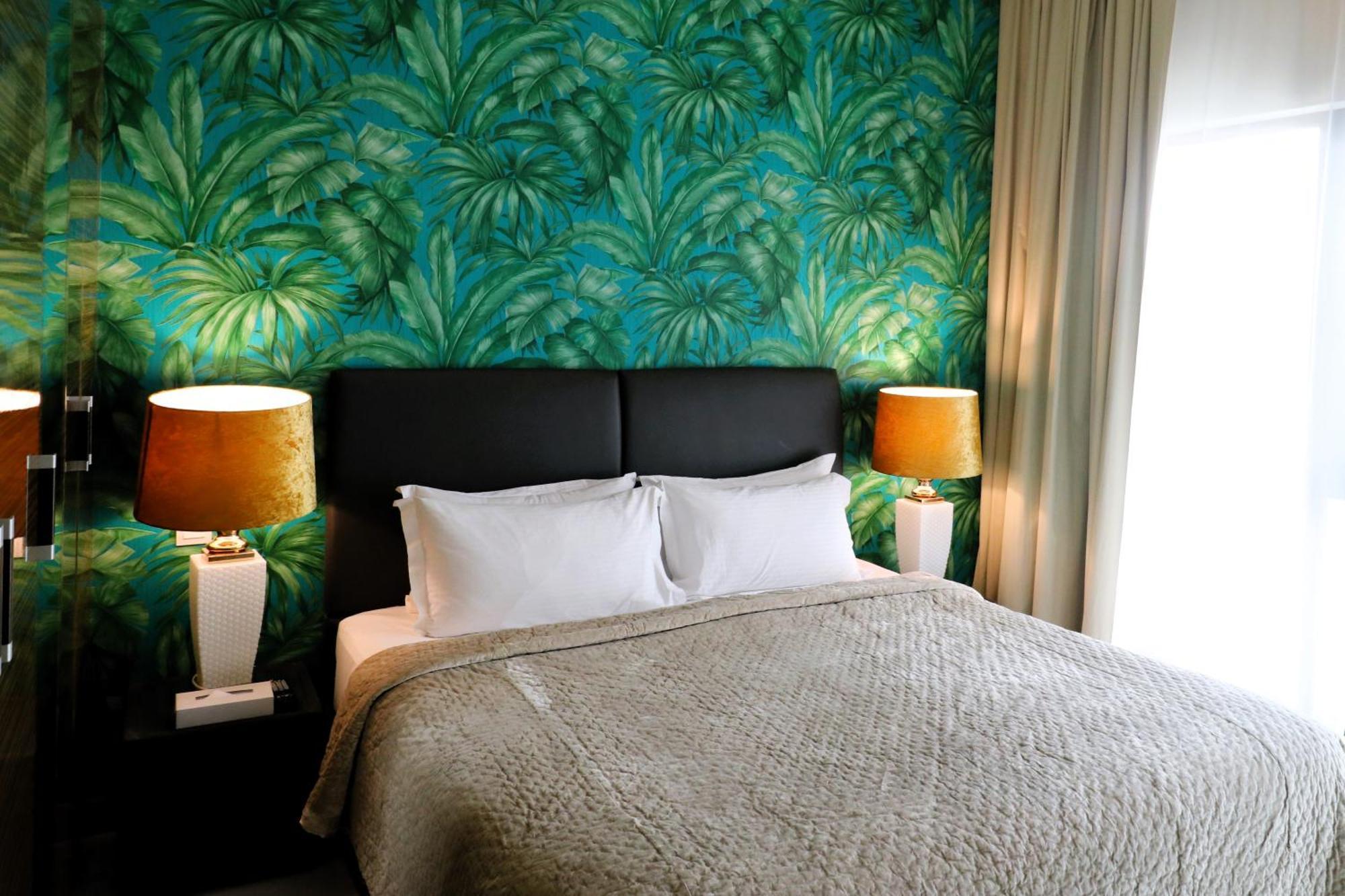 Lagoona Beach Luxury Resort And Spa Μανάμα Εξωτερικό φωτογραφία The photo shows a stylish bedroom featuring a large bed with white pillows and a gray blanket. The headboard is upholstered in black, providing a modern touch. Behind the bed, there is a vibrant green wallpaper with a tropical leaf pattern, adding a 