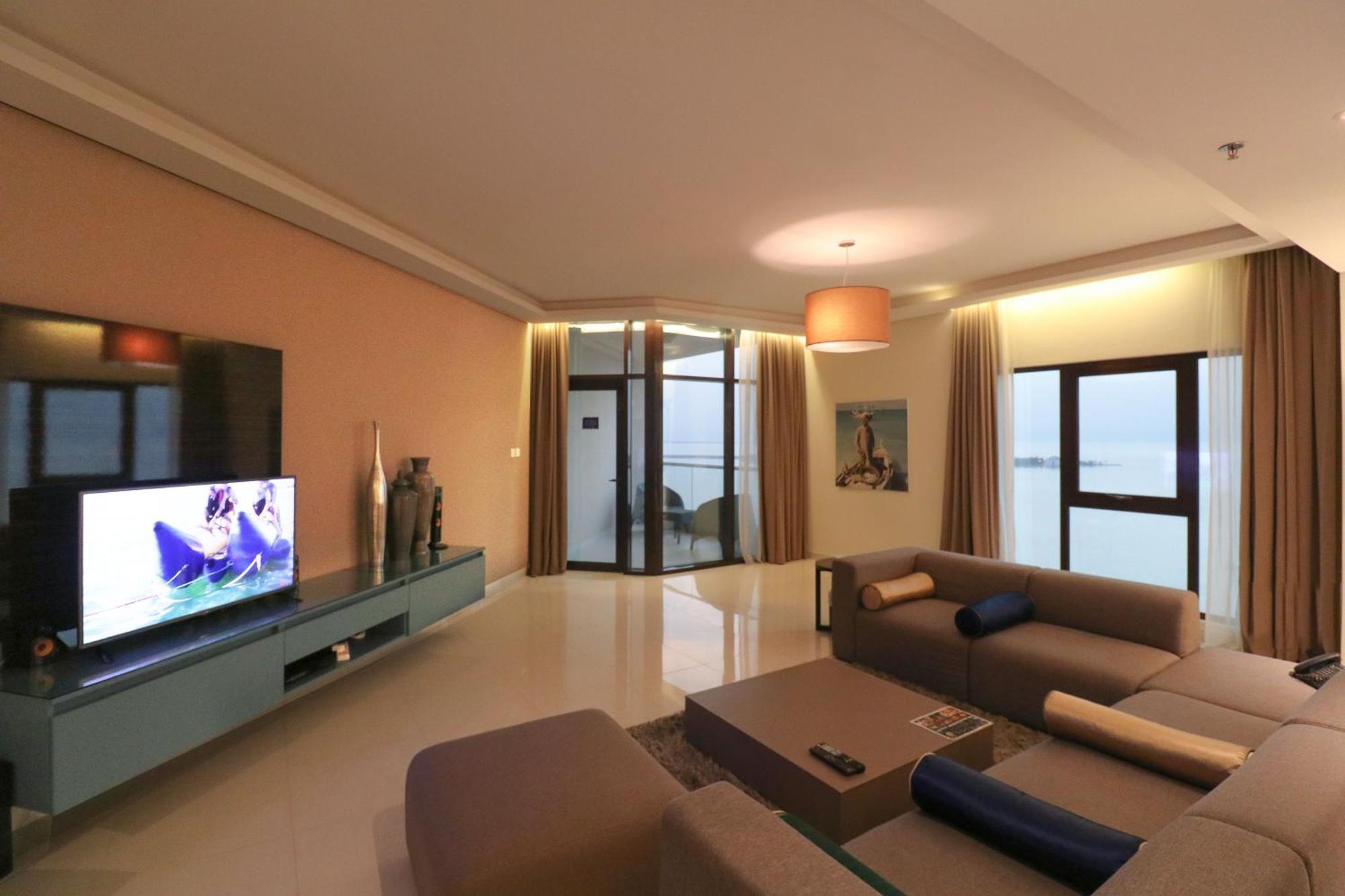 Lagoona Beach Luxury Resort And Spa Μανάμα Εξωτερικό φωτογραφία The photo shows a modern living room with a sleek design. There is a large wall-mounted television on a contemporary entertainment unit, with decorative items placed beside it. The room features a comfortable sectional sofa in light tones, with a cof