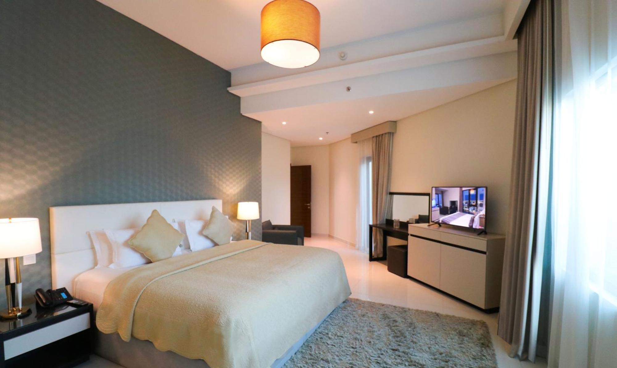Lagoona Beach Luxury Resort And Spa Μανάμα Εξωτερικό φωτογραφία The photo shows a modern hotel room featuring a large bed with a light-colored blanket and decorative pillows. The walls are painted in a subtle shade, and there is a textured accent wall behind the bed. On one side, a bedside lamp provides warm ligh