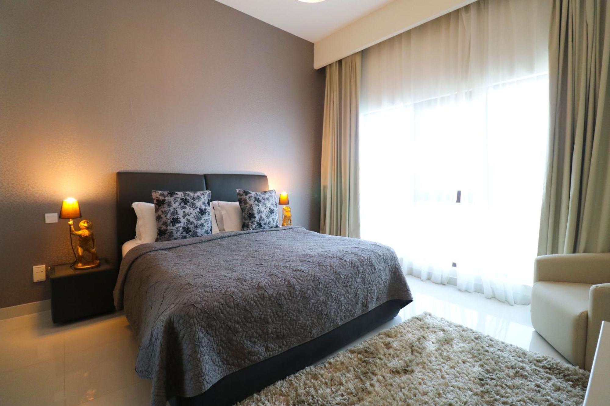 Lagoona Beach Luxury Resort And Spa Μανάμα Εξωτερικό φωτογραφία The photo shows a stylish bedroom with a modern design. It features a large bed with a dark gray bedspread and decorative pillows. On either side of the bed are two wooden nightstands, each with a lamp. There is a large window covered with light, flo